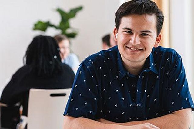 Ein Student der Uni Witten/Herdecke sitzt an einem Tisch und lächelt in die Kamera. Vor ihm liegen Lernunterlagen.