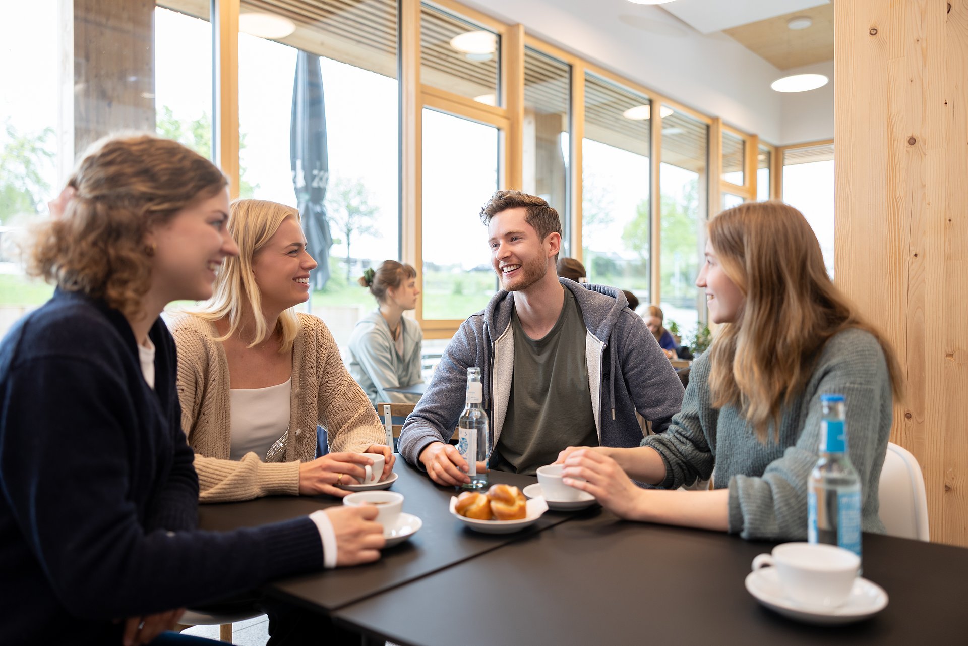 Eine Gruppe von vier jungen Menschen sitzt in einem Café, unterhält sich und trinkt Kaffee. 