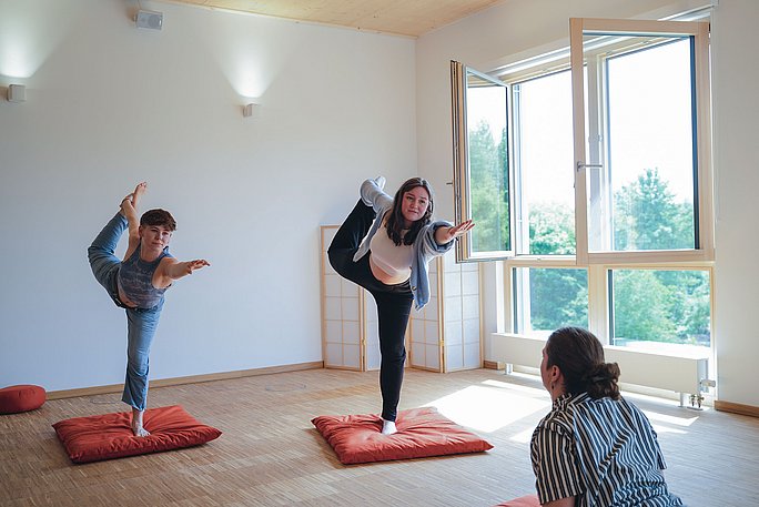 Im Fokus sind zwei Studentinnen, die die Yogapose "Tänzerin" einnehmen. Sie balancieren auf einem Bein und halten das andere hinter dem Rücken mit einer Hand fest, während der gegenüberliegende Arm nach vorne gestreckt ist.