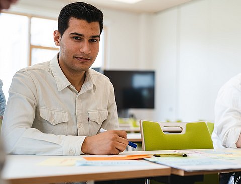 Ein junger Mann sitzt an einem Tisch in einem Seminarraum.