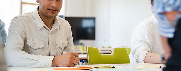 Ein junger Mann sitzt an einem Tisch in einem Seminarraum.