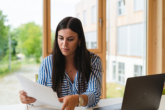 Eine Studentin sitzt vor ihrem Laptop und hält ein Blatt Papier in den Händen.