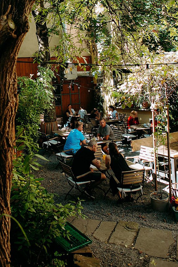 Blick in einen Biergarten: Am linken Bildrans ist ein Baum. In der Mitte sitzen Menschen an einer Holzsitzgruppe.