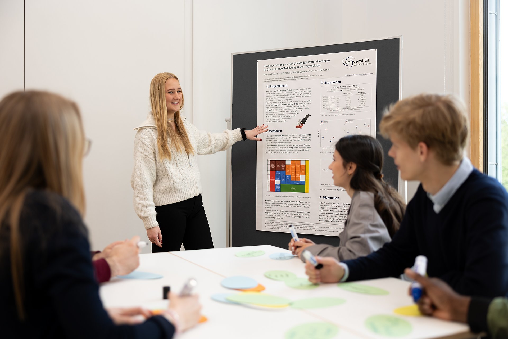 Eine Studentin zeigt auf ein Plakat, das an einer Metaplanwand hängt, und spricht zu einer Gruppe Studierender.