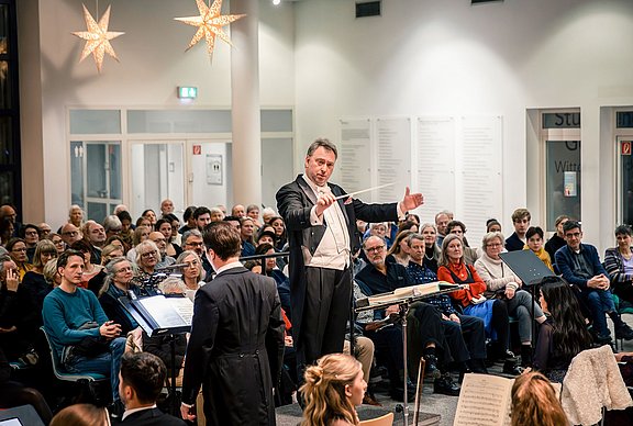 Ein Dirigent steht vor einem Orchester. Im Hintergrund sitzt das Publikum.