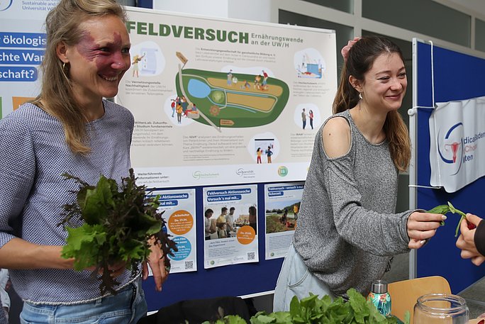 Zwei Frauen präsentieren ihr Projekt. Eine von ihnen hält einen Kopfsalat in den Händen. Hinter ihnen ist eine Pinnwand mit Infoplakaten.