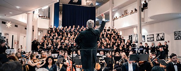 Foto eines Dirigenten der mit dem Rücken zum Publikum steht. Hinter ihm sitzt das Orchester und der Chor.