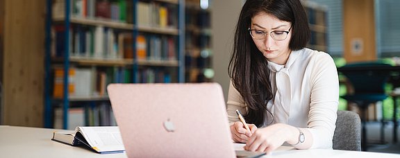 Eine Studentin sitzt an einem Schreibtisch und lernt. Vor ihr steht ein Laptop, daneben liegt ein aufgeschlagenes Buch. Im Hintergrund stehen Bücherregale.