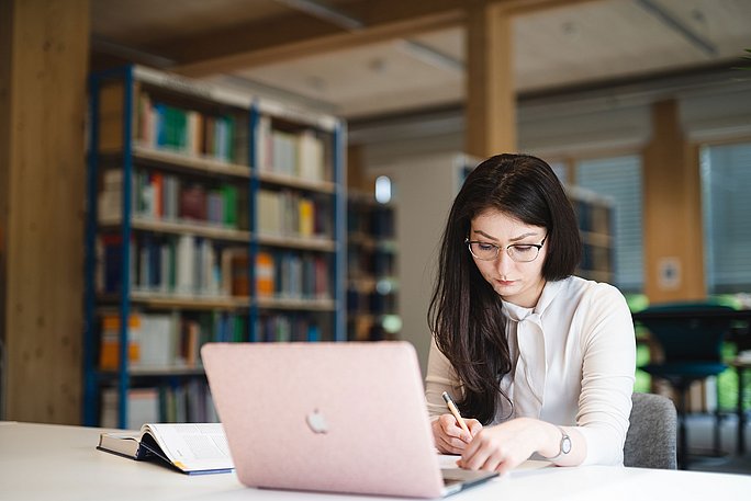 Eine Studentin sitzt an einem Schreibtisch und lernt. Vor ihr steht ein Laptop, daneben liegt ein aufgeschlagenes Buch. Im Hintergrund stehen Bücherregale.