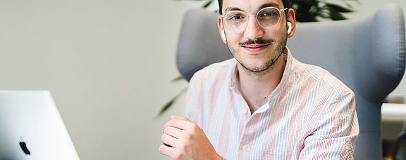 Student sitzt auf einem Sessel vor seinem Laptop und schaut in die Kamera.