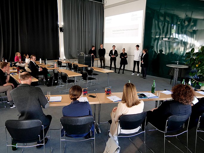 Foto von einem vollbesetzten Besprechungsraum. Vorne stehen Studierende, die eine Präsentation zeigen und ihr Projekt vorstellen.