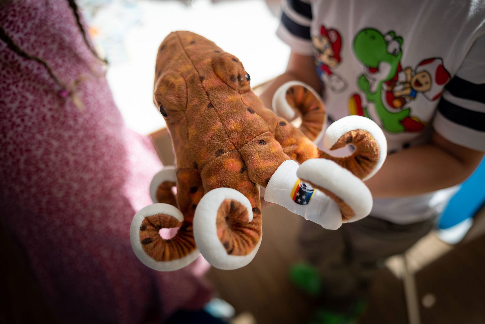 Im Fokus steht ein brauner Plüsch-OKtopus. Im Hintergrund sind Kinder zu erkennen, die mit ihm spielen.