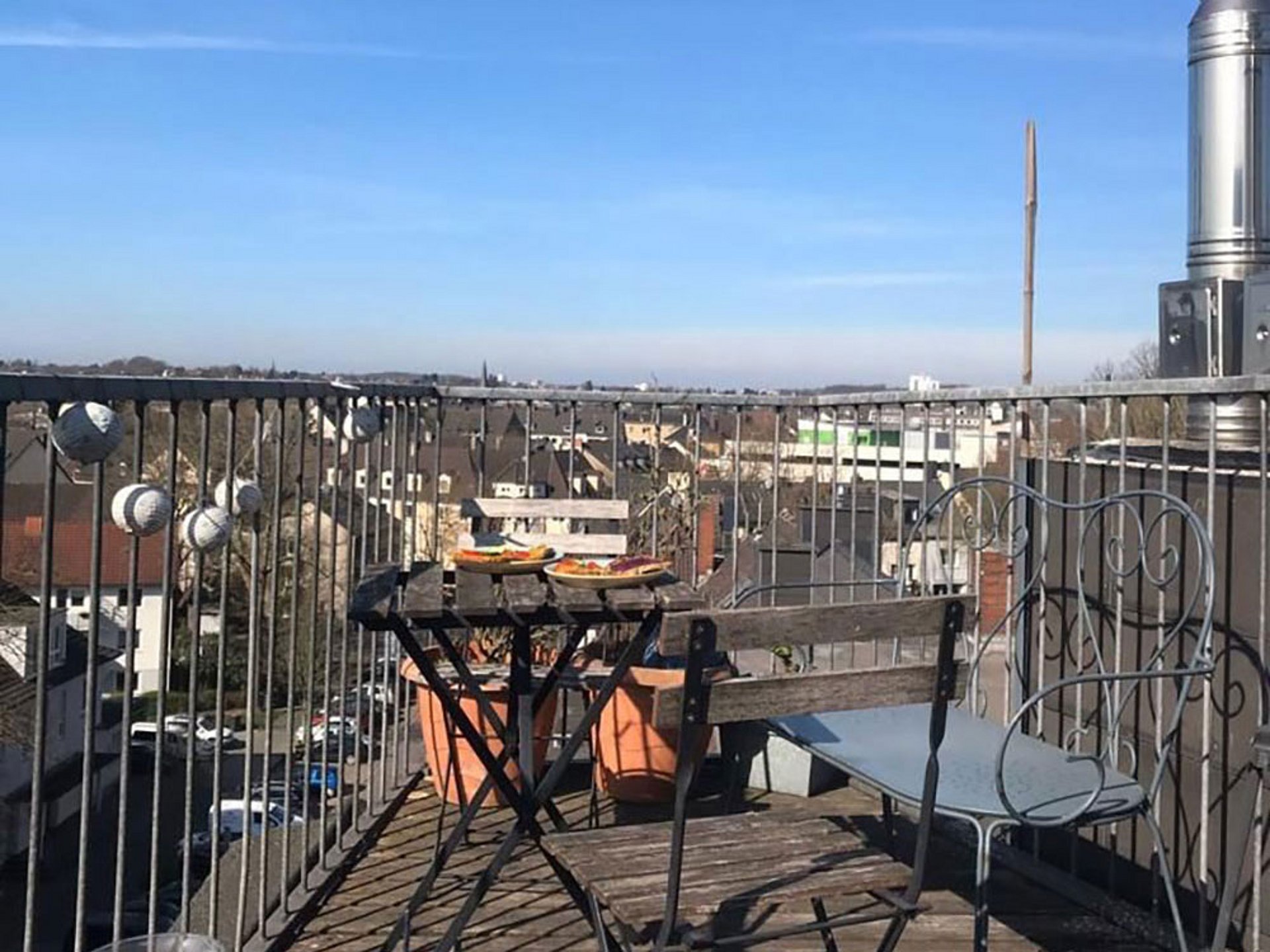 Auf einer kleinen Dachterrasse stehen zwei Holzklappstühle, eine Bank und ein Tisch. Die Terrasse ist von einem Metallgeländer umgeben. Die Hälfte des Fotos zeigt strahlendblauen Himmel.