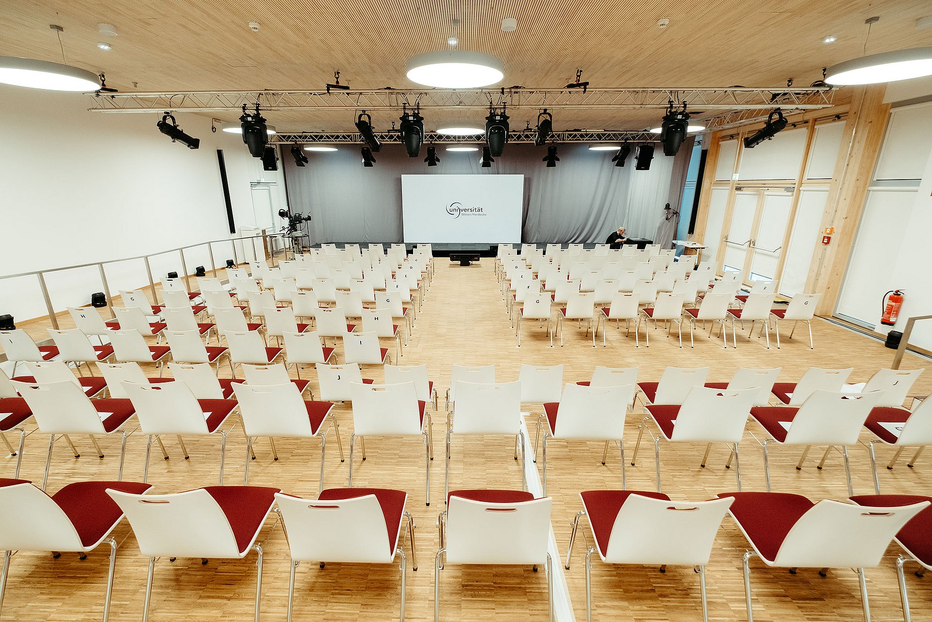 Shot of a hall in which red upholstered chairs with white backrests are arranged in rows, with a screen at the front