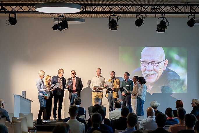 Eine Gruppe von Menschen steht auf einer Bühne und singt. Hinter ihnen wird das Foto eines Mannes an die Wand projeziert.