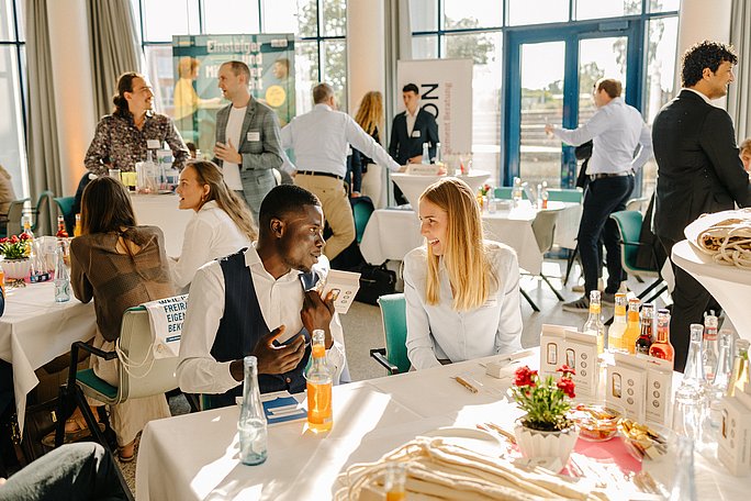 Foto von zwei Personen, die an einem Tisch sitzen und sich unterhalten. Im Hintergrund stehen weitere Menschen.