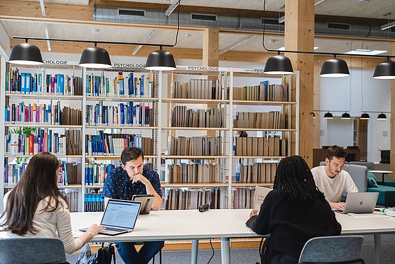 Vier Studierende sitzen an einem langen Tisch mit ihren Laptops und Büchern, um zu lernen.