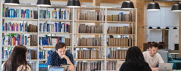 Vier Studierende sitzen an einem langen Tisch mit ihren Laptops und Büchern, um zu lernen.