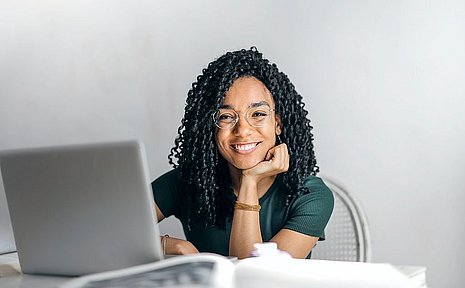 Eine junge Frau sitzt vor einem Laptop und lächelt in die Kamera.