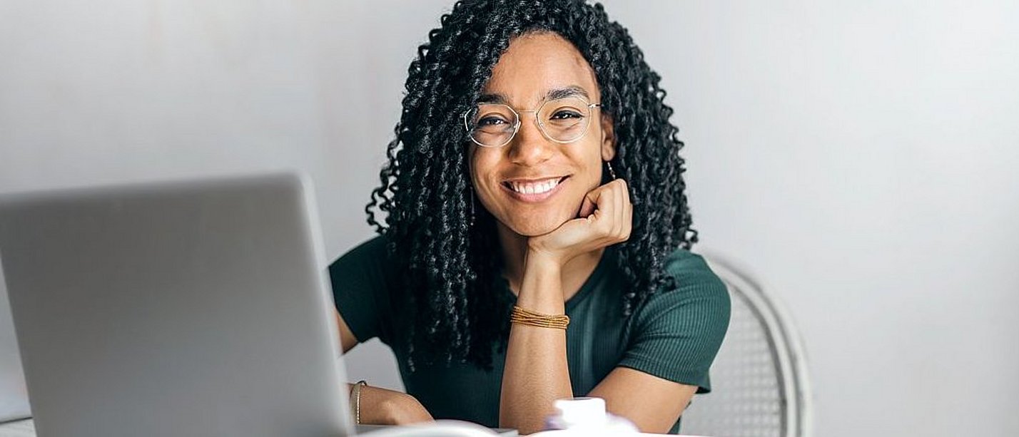 A young woman sits in front of a laptop and smiles at the camera.