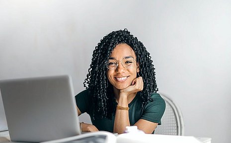 Eine junge Frau sitzt vor einem Laptop und lächelt in die Kamera.