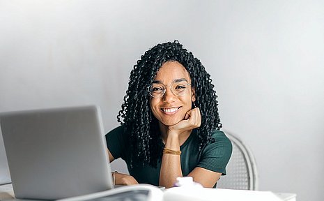 A young woman sits in front of a laptop and smiles at the camera.