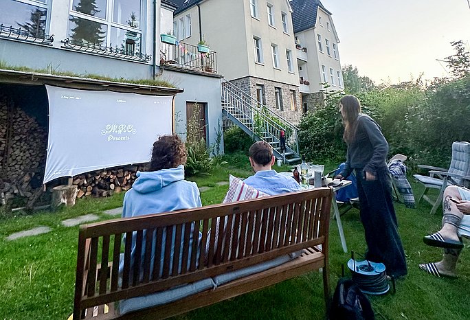 Auf einer Gartenbank sitzen zwei Studenten. Sie schauen auf eine Leinwand, die an einer Hauswand aufgespannt ist.