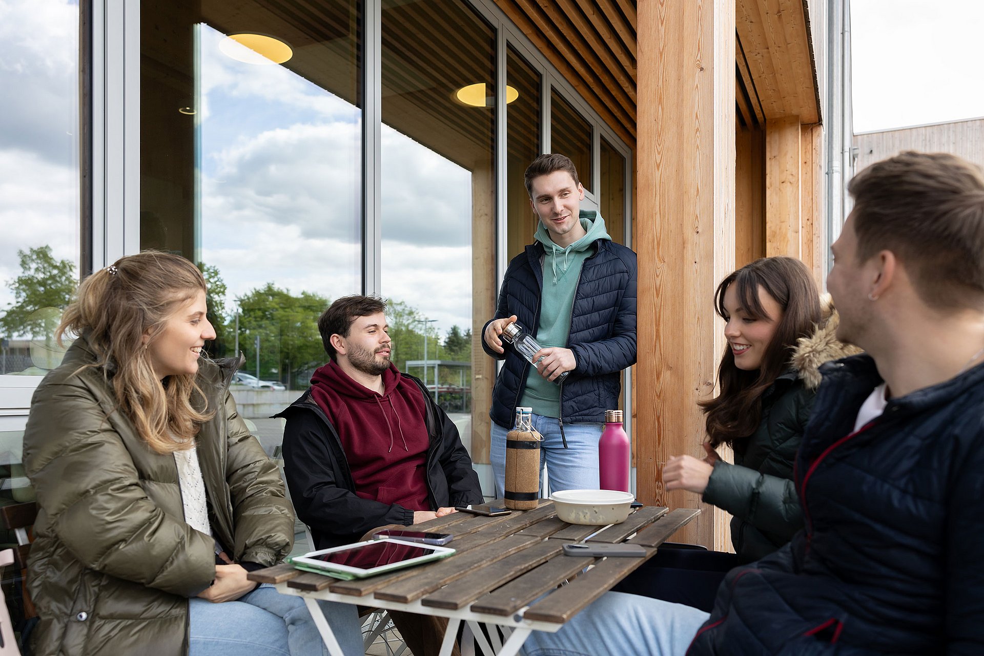 Eine Gruppe Studierender sitzt an einer Terrassen-Sitzgelegenheit vor dem Uni-Café und unterhält sich.