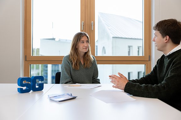 Eine Frau und ein Mann sitzen sich gegenüber. Hinter ihnen ist ein großes Fenster. Auf dem Tisch zwischen ihnen stehen die Buchstaben "S" und "G" als Aufsteller, dazwischen liegen Unterlagen.