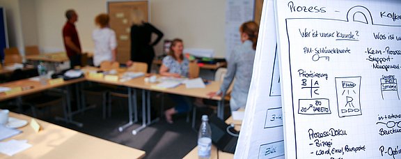 Flipchart mit Grafiken im Vordergrund, im Hintergrund reden fünf Personen.