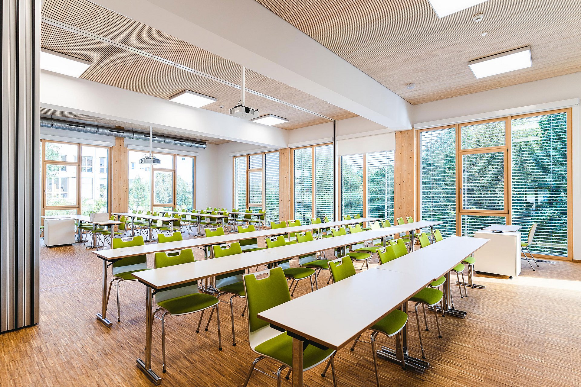 Rows of tables and chairs stand in a light-flooded room with a wooden floor and ceiling