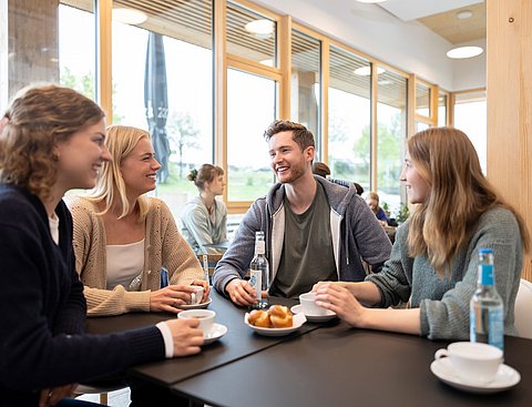 Vier Studierende sitzen gemeinsam an einem Tisch im Uni-Café. Vor ihnen stehen Kaffeetassen und Gebäck.