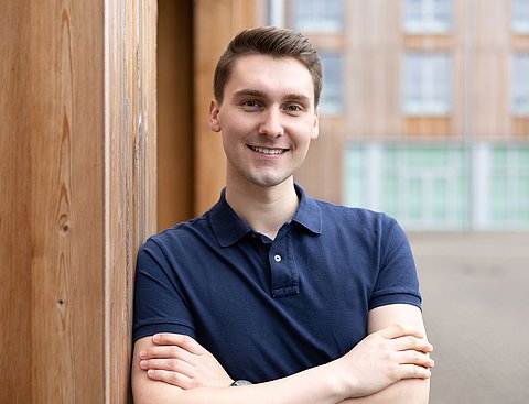 Ein junger Mann steht seitlich gelehnt an einer Gebäudewand aus Holz.