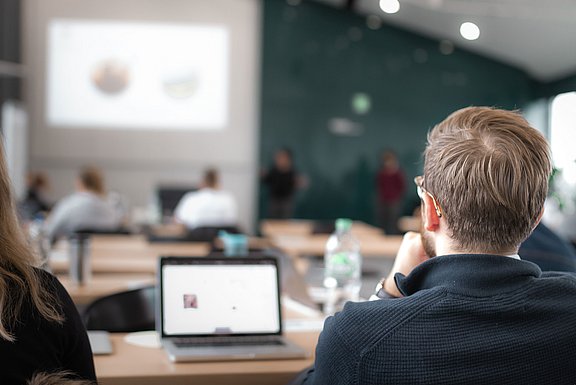 Blick über die Schulter eines Mannes, der auf eine Leinwand schaut und vor sich einen Laptop aufgestellt hat.