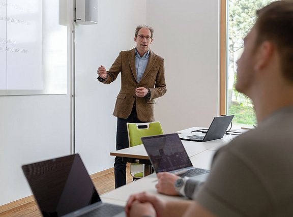 Ein Professor steht an einer Tafel und erklärte Studierenden etwas.