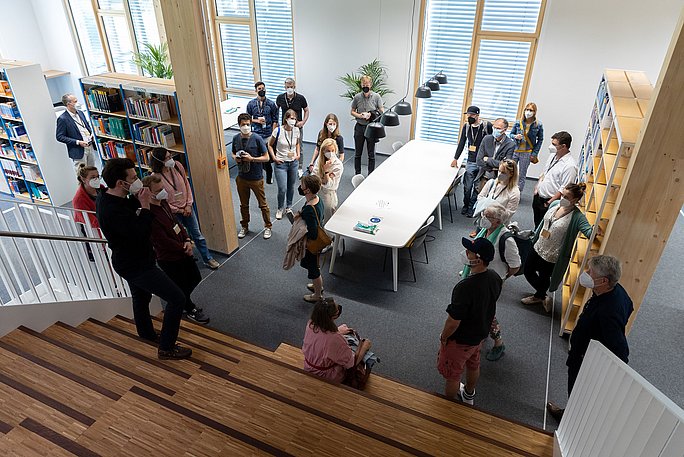 Eine Gruppe von Menschen steht in einer Bibliothek. Im Vordergrund ist ein Treppenabgang.