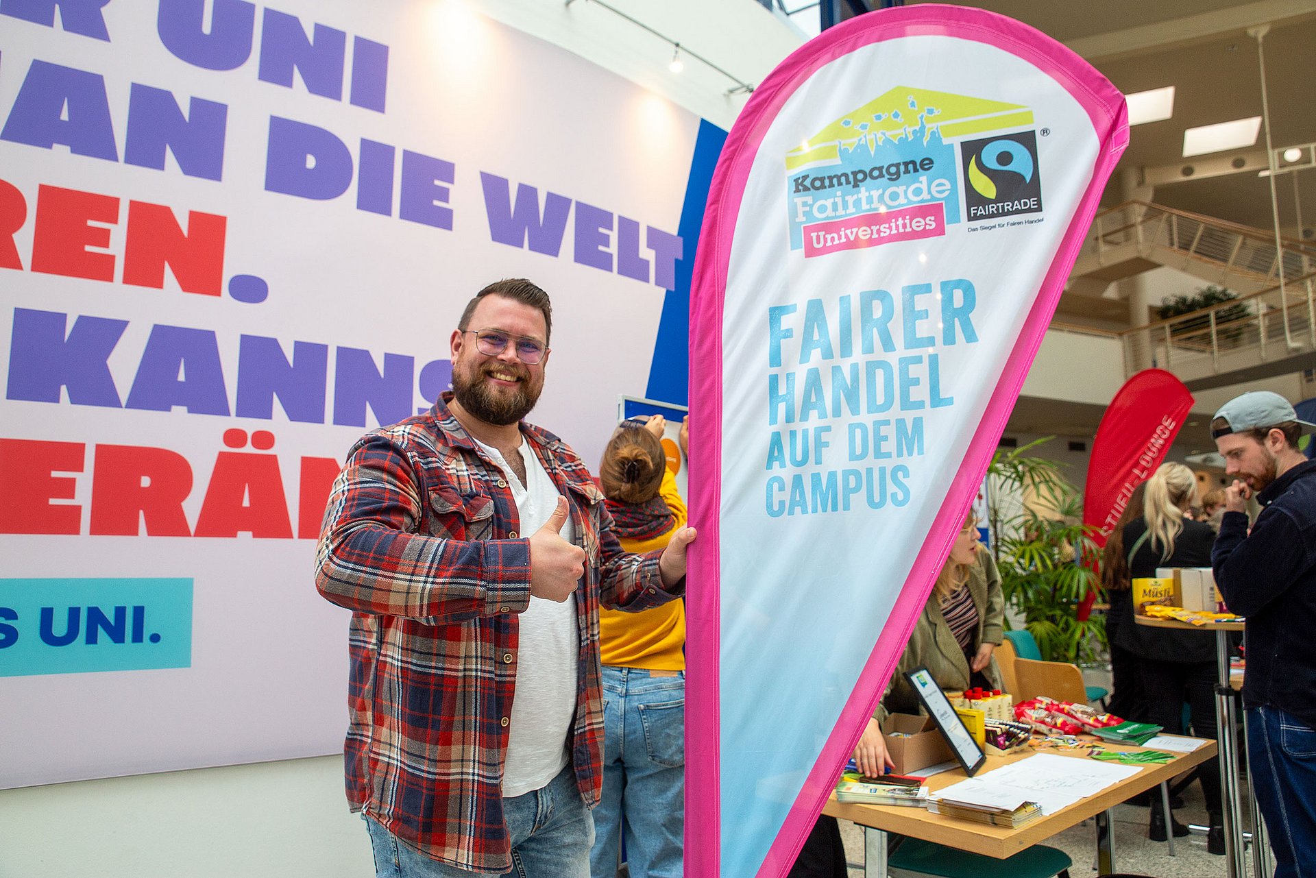 Ein Mann lächelt mit nach oben gestreckten Daumen in die Kamera. Er steht neben einer Beachflag, die das Siegel "Fairtrade University" ziert.