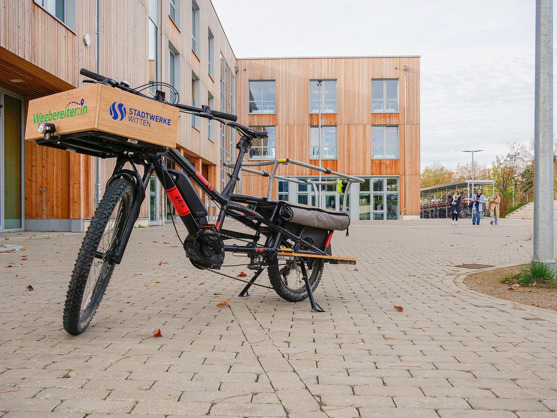 Im Mittelpunkt des Fotos steht ein Lastenfahrrad. Im Hintergrund ist die Holzfassade des Neubaus der Uni zu sehen.