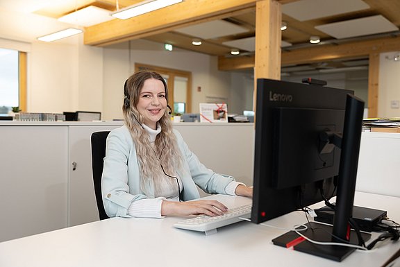 Eine junge Frau mit Headset sitzt vor einem Bildschirm und lächelt in die Kamera.