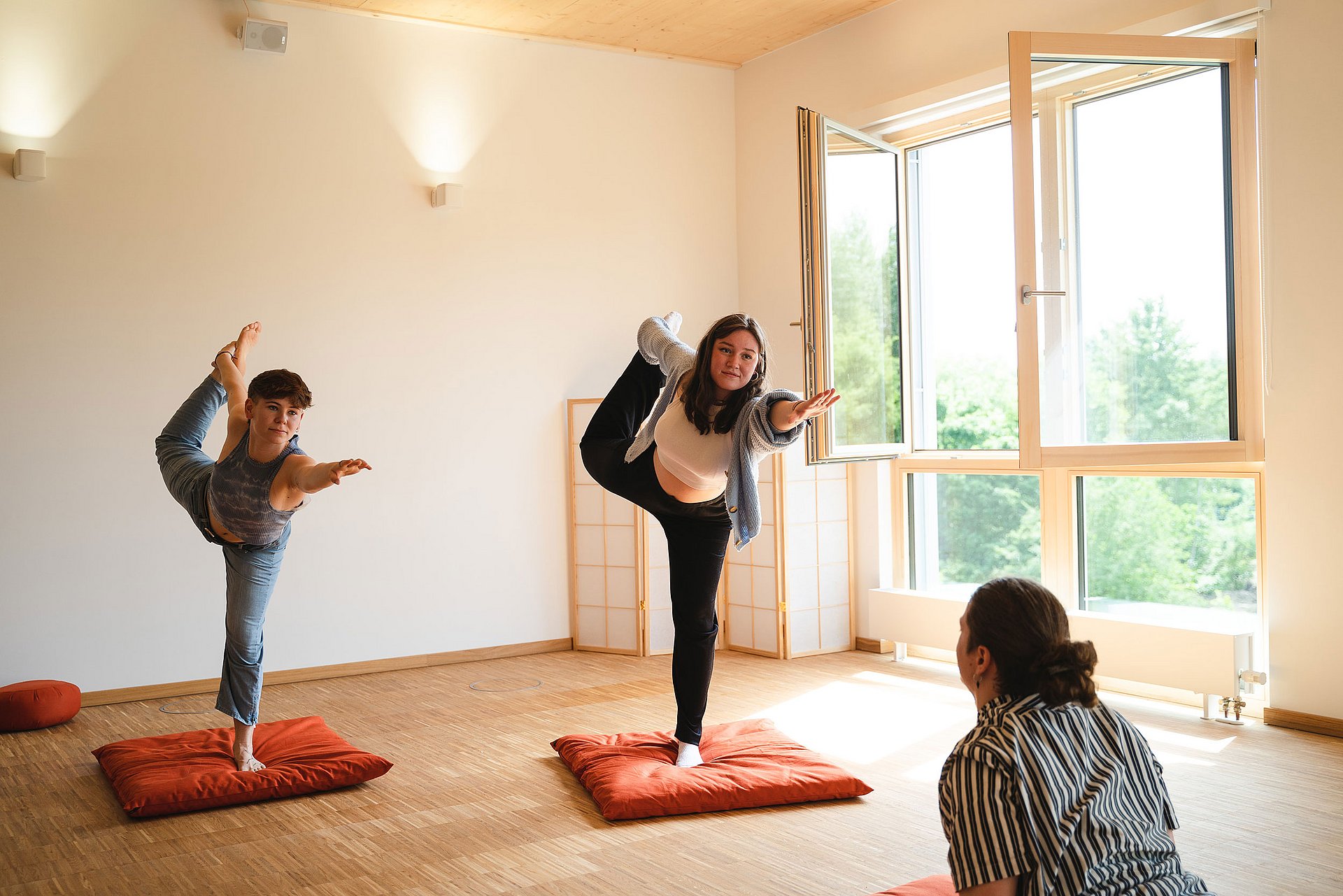 Two students stand with their left leg on a mat, pull their right leg back into the air with their right arm and stretch their left arm forward.