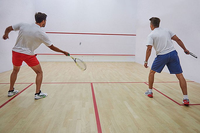 Zwei Männer spielen in einer Sporthalle Squash. Einer von ihnen schlägt gerade den Ball gegen die Wand.