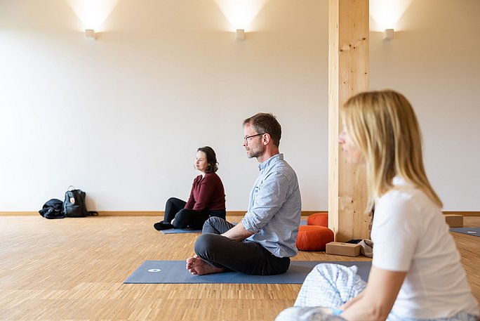 Drei Personen sitzen im Yoga-Sitz auf Sportmatten.