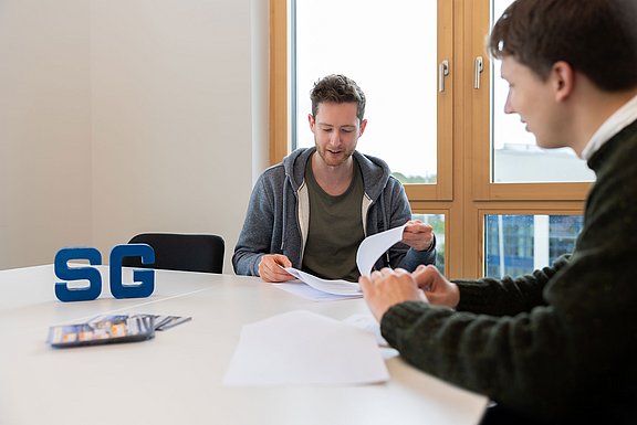 Ein Student sitzt zusammen mit einem Berater an einem Besprechungstisch. Sie schauen sich gemeinsam Vertragsunterlagen an. Auf dem Tisch steht ein Aufsteller der Studierenden-Gesellschaft.