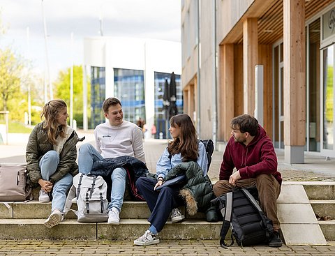 Eine Gruppe Studierender sitzt auf den Campustreppen neben dem Holzgebäude der Uni Witten/Herdecke. Sie sind in ein Gespräch vertieft.