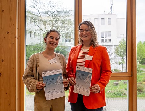 Foto von zwei Frauen, die nebeneinander vor einem Fenster stehen. Beide halten eine Urkunde in den Händen.