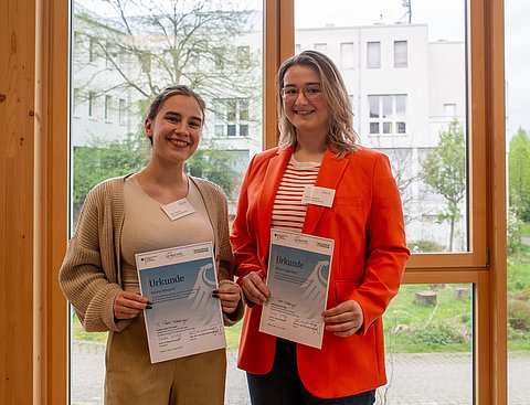 Foto von zwei Frauen, die nebeneinander vor einem Fenster stehen. Beide halten eine Urkunde in den Händen.