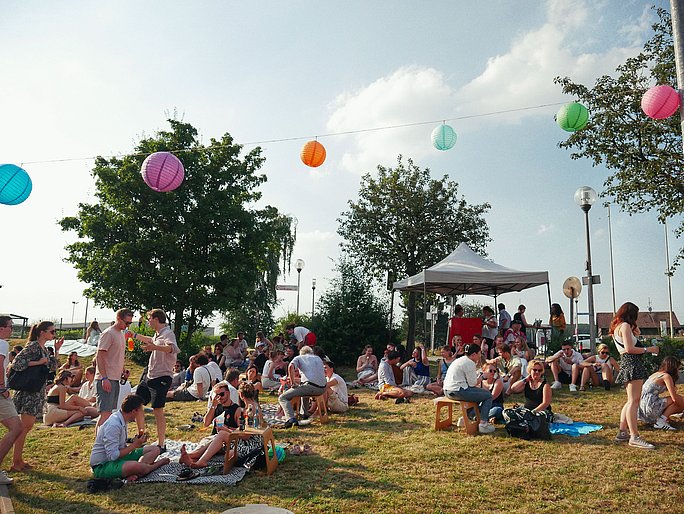 Menschen stehen oder sitzen auf einer Wiese. Über ihnen ist eine bunte Lampion-Girlande gespannt.