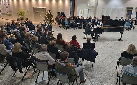 Menschen sitzen in einer großen Halle und hören einem Klavierspieler zu.