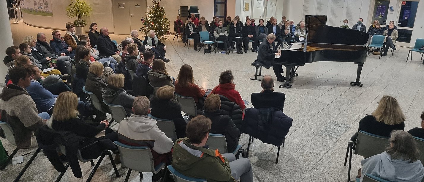 Menschen sitzen in einer großen Halle und hören einem Klavierspieler zu.