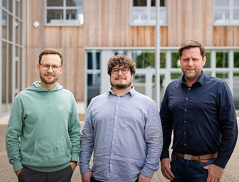 Foto von drei Männern, die nebeinander vor einem Gebäude mit Holzfassade stehen.